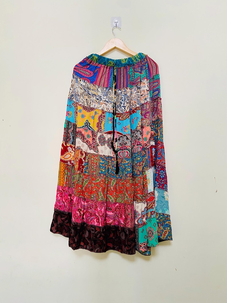 Puede incluir: Una falda larga de patchwork con una variedad de patrones coloridos, incluyendo dise&ntilde;os de cachemira y florales. La falda est&aacute; colgada en una percha de madera sobre un fondo blanco. La falda tiene un cord&oacute;n en la cintura y una borla negra.
