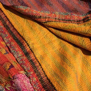 May include: Close-up of a colorful textile with a patchwork design. The fabric features a mix of orange, yellow, red, and blue hues, with intricate stitching and patterns. The texture appears soft and layered, suggesting a handcrafted item.