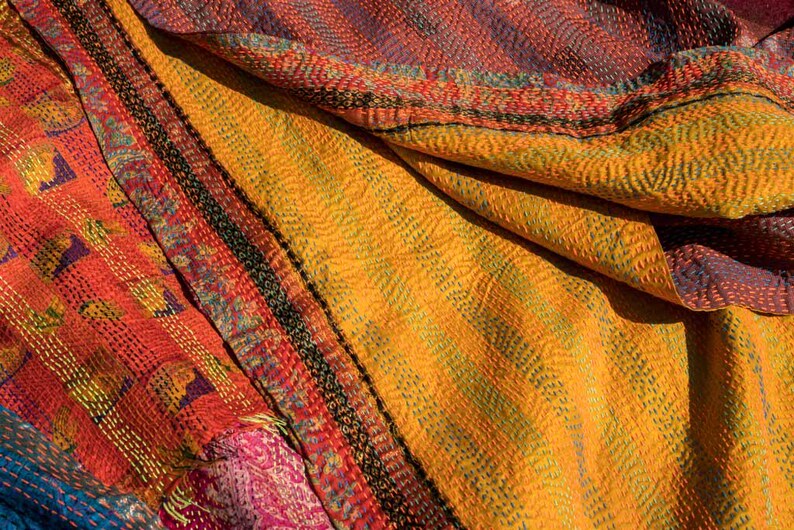 May include: Close-up of a colorful textile with a patchwork design. The fabric features a mix of orange, yellow, red, and blue hues, with intricate stitching and patterns. The texture appears soft and layered, suggesting a handcrafted item.