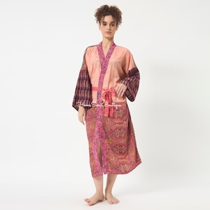 May include: A knee-length kimono robe featuring a peach-colored bodice with dark purple paisley patterned sleeves and a matching lower section. The garment includes a bright pink paisley trim along the front opening and a matching tie-waist belt.