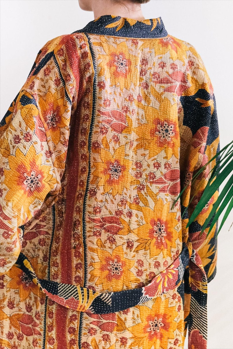 May include: A floral patterned kimono in shades of yellow, orange, and cream. The kimono features large flower designs and a contrasting navy blue trim. The belt is patterned with stripes and floral accents. The garment is made of a soft, quilted fabric.