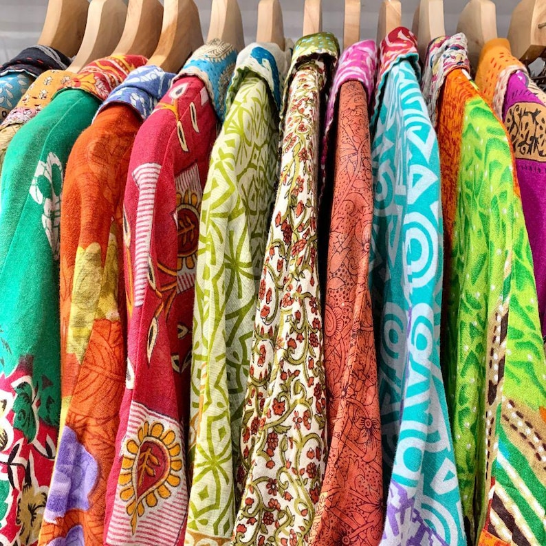 K&ouml;nnte beinhalten: Eine Sammlung farbenfroher, gemusterter Hemden, die an Holzb&uuml;geln h&auml;ngen. Die Hemden zeigen eine Vielzahl lebendiger Farben, darunter Rot, Orange, Gr&uuml;n und Blau, mit aufwendigen Blumen- und geometrischen Designs. Die Stoffe wirken leicht und flie&szlig;end.