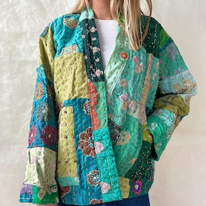 Könnte beinhalten: Eine Patchwork-Jacke in Türkis, Grün und Gold. Die Jacke hat verschiedene Blumen- und geometrische Muster mit sichtbaren Nähten. Die Ärmel sind lang und die Jacke hat einen entspannten, offenen Schnitt.