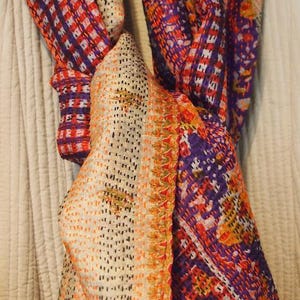 May include: A colorful scarf with a patchwork design. The scarf features a mix of orange, purple, and white patterns. The fabric appears to be soft and lightweight, suitable for various seasons. The scarf is draped, showcasing its texture and design.