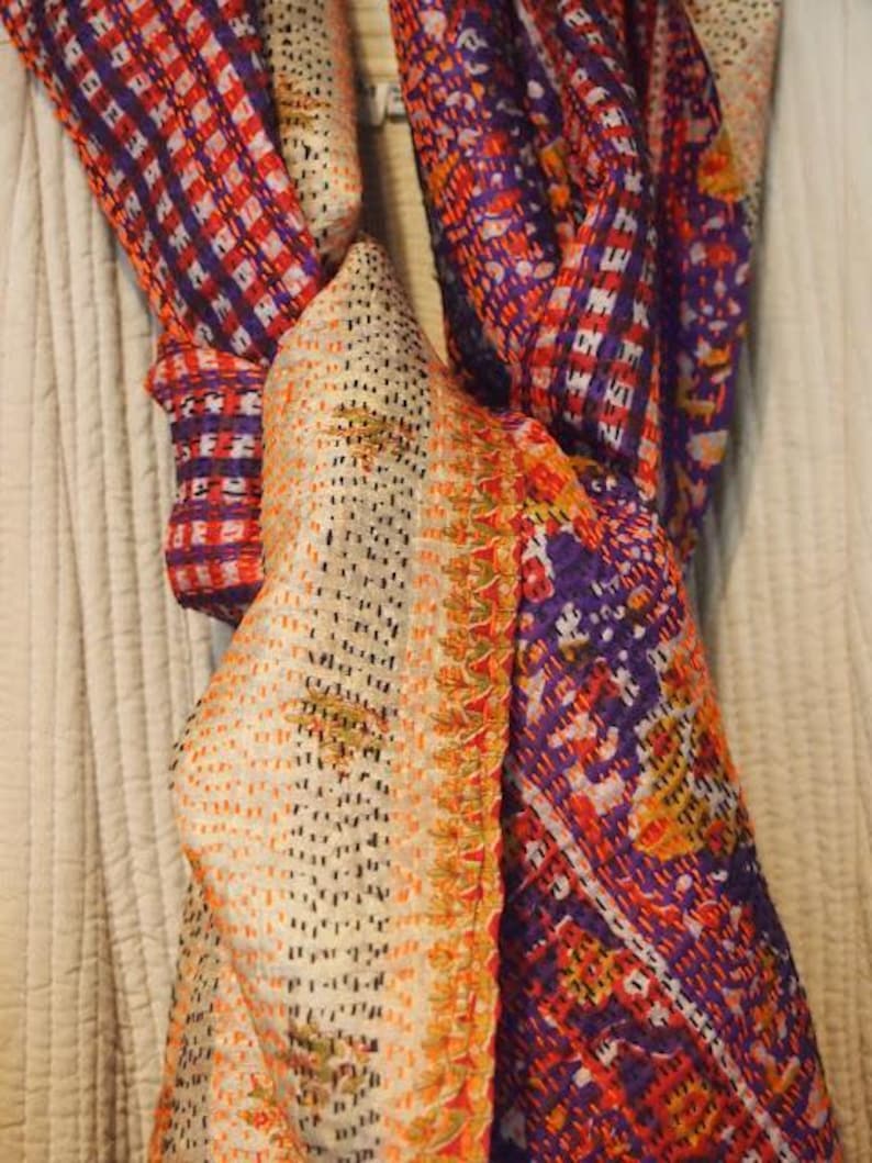 May include: A colorful scarf with a patchwork design. The scarf features a mix of orange, purple, and white patterns. The fabric appears to be soft and lightweight, suitable for various seasons. The scarf is draped, showcasing its texture and design.