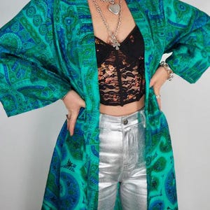 K&ouml;nnte beinhalten: Ein langer, flie&szlig;ender Kimono mit t&uuml;rkisblauem und blauem Paisley-Muster. Der Kimono wird mit einem schwarzen Spitzentop und einer silberfarbenen Metallic-Hose kombiniert. Das Outfit wird mit mehrschichtigen Silberketten und einem Armband erg&auml;nzt.