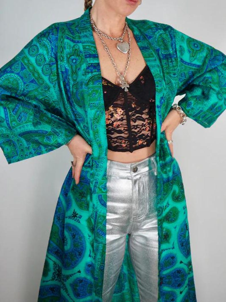 K&ouml;nnte beinhalten: Ein langer, flie&szlig;ender Kimono mit t&uuml;rkisblauem und blauem Paisley-Muster. Der Kimono wird mit einem schwarzen Spitzentop und einer silberfarbenen Metallic-Hose kombiniert. Das Outfit wird mit mehrschichtigen Silberketten und einem Armband erg&auml;nzt.