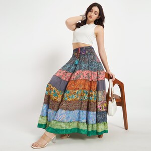 May include: A colorful patchwork maxi skirt with a drawstring waist. The skirt features a variety of patterned fabrics in shades of blue, pink, orange, and green. The model is wearing a white crop top and holding a white purse.
