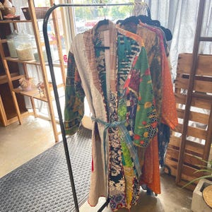 May include: A colorful patchwork robe with a blue tie, hanging on a black rack. The robe features a variety of patterns and colors, including green, red, and floral designs. Other robes in various colors are also visible.