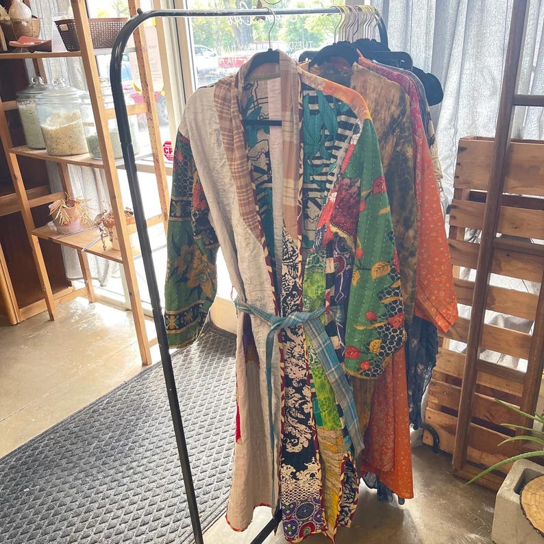 May include: A colorful patchwork robe with a blue tie, hanging on a black rack. The robe features a variety of patterns and colors, including green, red, and floral designs. Other robes in various colors are also visible.