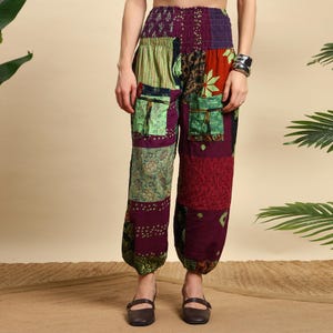 May include: Patchwork pants in shades of green, purple, and red. The pants feature a mix of patterned fabrics, including floral and striped designs. Two pockets are visible on the front. The pants have an elastic waistband and are worn with brown shoes.