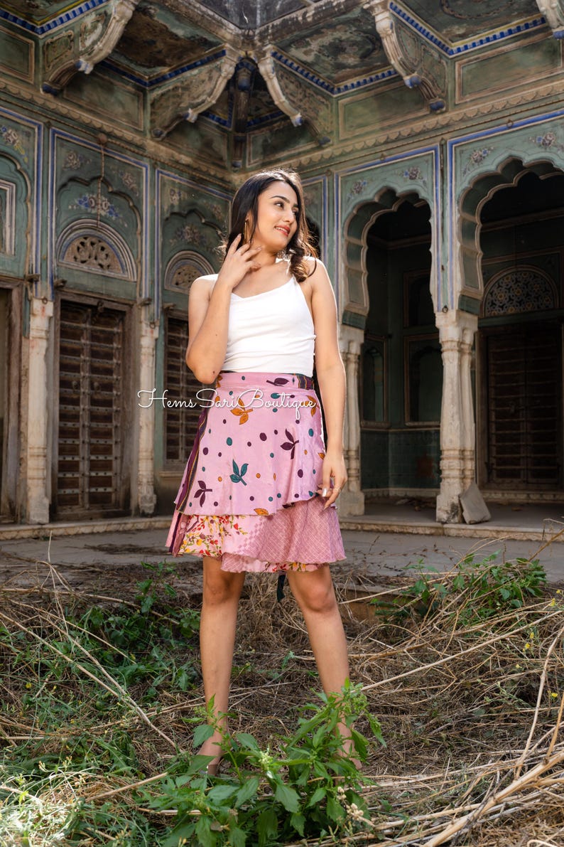 May include: A layered pink skirt with a floral and leaf pattern, paired with a white tank top. The skirt features a wrap design and is adorned with colorful accents. The image also shows the text "Hems Sari Boutique".