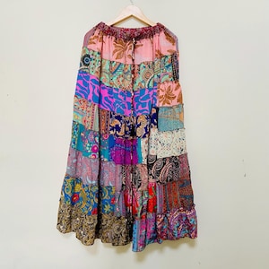 Puede incluir: Falda patchwork con una variedad de patrones coloridos, incluyendo dise&ntilde;os de cachemira y florales. La falda est&aacute; hecha de m&uacute;ltiples paneles de tela y tiene un dobladillo con volantes. Cuelga de una percha de madera contra una pared neutra.