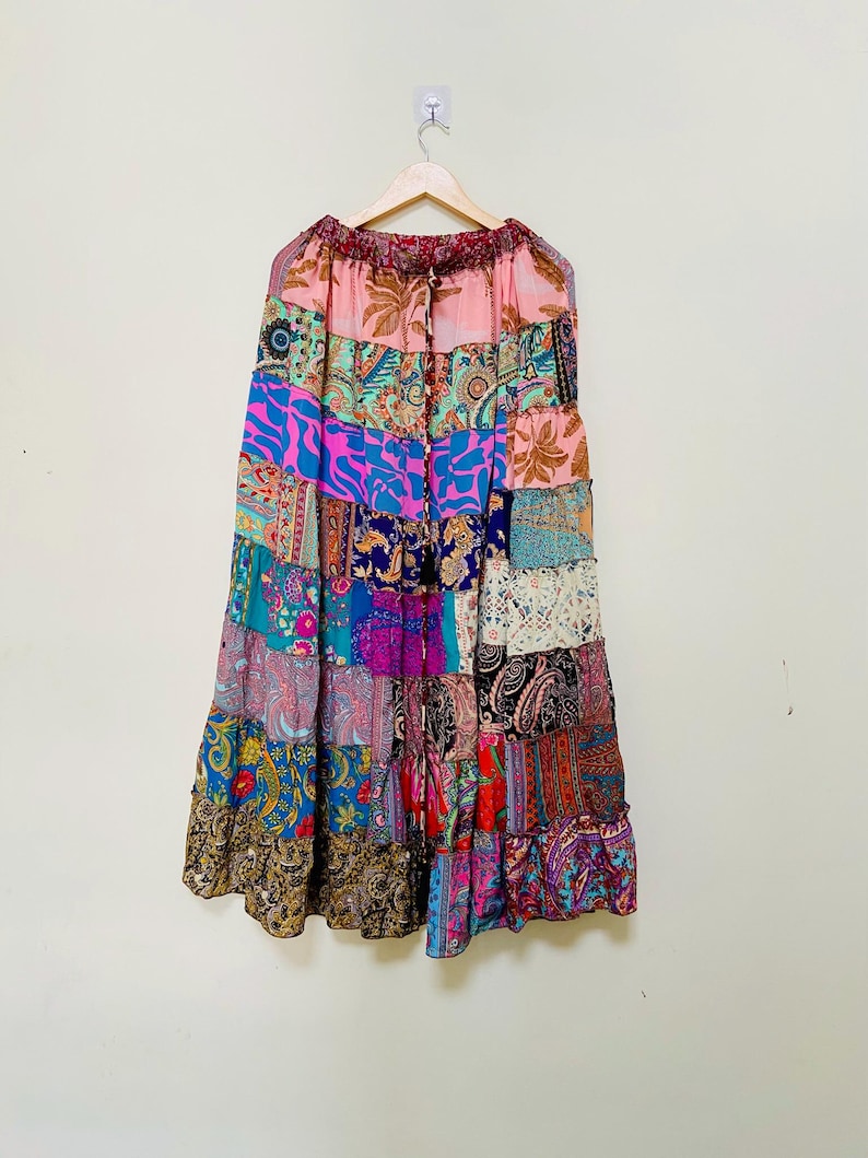 Puede incluir: Falda patchwork con una variedad de patrones coloridos, incluyendo dise&ntilde;os de cachemira y florales. La falda est&aacute; hecha de m&uacute;ltiples paneles de tela y tiene un dobladillo con volantes. Cuelga de una percha de madera contra una pared neutra.