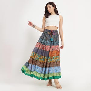 May include: A long, tiered skirt with a patchwork design in various colors, including blue, pink, orange, and green. The skirt has a drawstring waist and a ruffled hem. The model is wearing a white crop top.