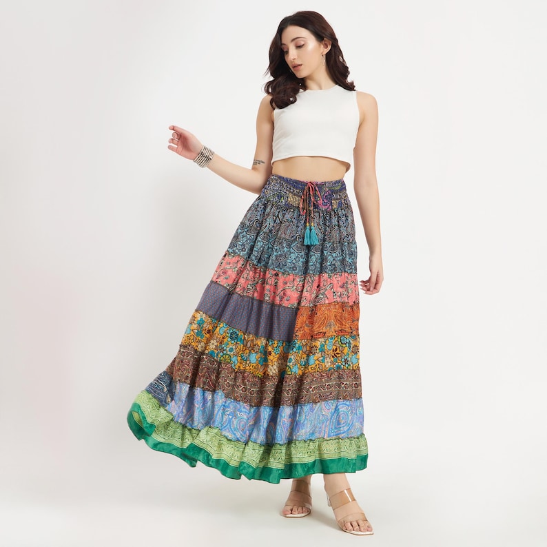 May include: A long, tiered skirt with a patchwork design in various colors, including blue, pink, orange, and green. The skirt has a drawstring waist and a ruffled hem. The model is wearing a white crop top.