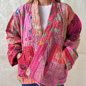 Könnte beinhalten: Eine Patchwork-Jacke in Rosa-, Korallen- und Fuchsiatönen. Die Jacke hat aufwendige Stickereien und Nähte mit einem entspannten, offenen Design. Die Ärmel sind lang und die Jacke hat zwei Vordertaschen. Das Kleidungsstück wird über einem weißen Oberteil und einer blauen Jeans getragen.