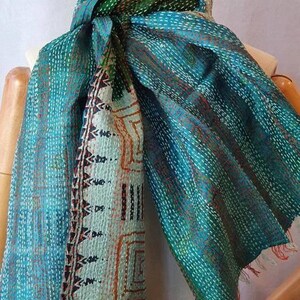 May include: A teal and green scarf with intricate stitching and a patterned section. The scarf has a fringed edge and is draped over a mannequin. The design includes geometric and abstract patterns.