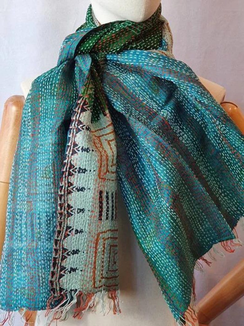 May include: A teal and green scarf with intricate stitching and a patterned section. The scarf has a fringed edge and is draped over a mannequin. The design includes geometric and abstract patterns.