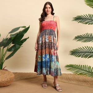 Op de afbeelding: Een patchwork midi-jurk met een rode, getextureerde lijfje en een patroonrok. De rok heeft vierkanten in verschillende kleuren en ontwerpen, waaronder blauw, bruin en teal. De jurk heeft dunne bandjes en een vloeiend silhouet, geschikt voor warm weer.