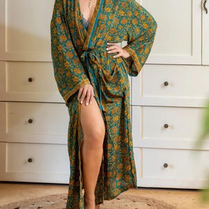 K&ouml;nnte beinhalten: Ein langer, flie&szlig;ender Bademantel mit einem teal- und goldfarbenen Blumenmuster. Der Bademantel hat weite &Auml;rmel, einen Taillenbindeg&uuml;rtel und einen hohen Schlitz. Das Kleidungsst&uuml;ck wird offen getragen und gibt einen Blick auf ein blaues Unterkleid frei.