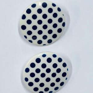 May include: Two round, white earrings with a navy blue polka dot pattern. The earrings are set against a white background, showcasing their glossy finish and symmetrical design. These earrings are a classic accessory.