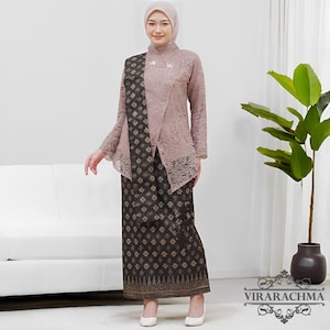 May include: A traditional Indonesian outfit. The outfit features a blush pink lace top with long sleeves and a matching head covering. The outfit includes a dark brown skirt with a gold geometric pattern and a matching shawl. The model is wearing white heels.