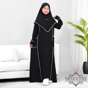 Può includere: Un abito abaya nero con finiture rosa chiaro e hijab abbinato. L'abito presenta maniche lunghe, tasche e un design modesto. L'hijab incornicia il viso e l'outfit è adatto a occasioni religiose o culturali.