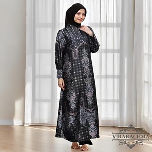 May include: A long, black dress with a detailed floral pattern in shades of gray and pink. The dress has long sleeves and a high neckline. The model is wearing a black head covering. The dress is floor-length and has a flowing silhouette.