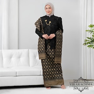 Op de afbeelding: Een vrouw draagt een zwart traditioneel kostuum met gouden accenten. De outfit bestaat uit een top met lange mouwen, een rok en een sjaal. Een gouden broche siert de top. De rok en sjaal hebben een herhalend geometrisch patroon. De outfit wordt gedragen met een hoofdbedekking.