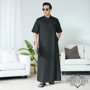May include: A black, short-sleeved Thobe, a traditional Arab garment, is worn by a person. The Thobe features a mandarin collar, a chest pocket, and side pockets. The person is wearing black sandals and sunglasses. The brand name "VIRARACHMA" is visible at the bottom.