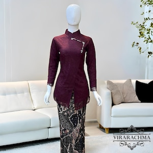 Puede incluir: Una blusa de encaje burdeos con mangas largas y cuello mandarín, combinada con una falda estampada. La blusa presenta un adorno decorativo cerca del cuello. La falda tiene un diseño floral negro y marrón. El conjunto se exhibe en un maniquí.