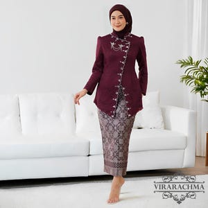 May include: A burgundy traditional outfit, featuring a long-sleeved jacket with intricate silver embellishments and a matching patterned skirt. The jacket has a mandarin collar and a decorative closure. The skirt has a detailed, repeating pattern. The outfit is worn with a matching head covering.