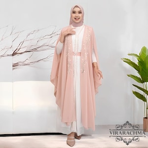 May include: A woman wearing a white dress with a light pink cape and a matching head covering. The cape is adorned with floral embroidery and a belt. The outfit is elegant and suitable for special occasions.