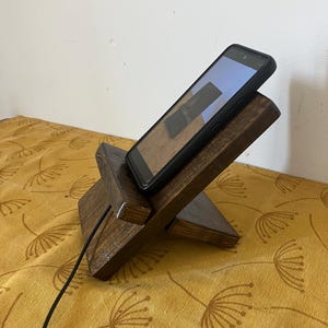 Peut inclure: Un support de téléphone en bois brun foncé avec un téléphone dans un étui noir. Le support est incliné pour la visualisation et dispose d'une fente pour un câble de chargement. Le téléphone affiche une image. Le support est sur une surface jaune.