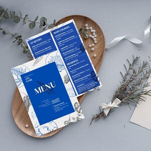 May include: A restaurant menu with a blue and white nautical design. The menu includes starters, main courses, and desserts. The menu is presented on a wooden tray with decorative elements, including pearls and a sprig of rosemary.