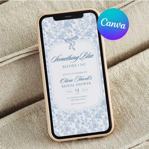 Puede incluir: Un smartphone muestra una invitación a una despedida de soltera con el texto "Something Blue". La invitación presenta un diseño floral en azul claro y blanco. El teléfono tiene un marco dorado y está sobre una tela beige.