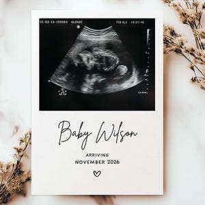 May include: A pregnancy announcement card with a sonogram image at the top. The text "Baby Wilson" is written in elegant script, with "Arriving November 2026" below, and a heart symbol at the bottom. The card is on a neutral background.