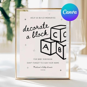 Könnte beinhalten: Ein gerahmtes Schild mit goldenem Rand zeigt die Worte "decorate a block" über einer Grafik von Spielzeugblöcken. Das Schild enthält den Text "Help us build memories" und "For Baby Robinson". Ein Canva-Logo befindet sich in der oberen rechten Ecke.