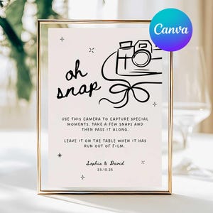 May include: A framed sign with the words "oh snap" in black script, featuring a line drawing of a camera and text. The sign includes instructions for capturing moments and is set against a blurred background.