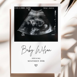 May include: A black and white sonogram image on a white card. The card reads "Baby Wilson Arriving November 2026" in elegant script, with a small heart symbol below the text. The image is a pregnancy announcement.