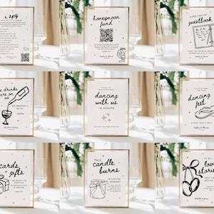 May include: A collection of framed signs with gold borders, featuring black text and illustrations on a cream background. The signs include phrases like "I Spy," "Honeymoon Fund," "Guestbook," "Dancing with Us," and "Cards + Gifts," suitable for a wedding or event.