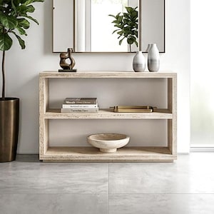 May include: A rectangular console table made of light-colored stone with two open shelves, featuring a square mirror above it, a tall potted plant in a bronze-toned planter, two ceramic vases, a small abstract sculpture, and a stone bowl.