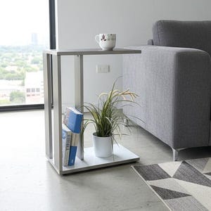 Könnte beinhalten: Ein moderner Beistelltisch mit grauem Oberteil und weißer Basis, mit einem eingebauten Regal mit Büchern und einer Topfpflanze. Eine Tasse steht auf der Tischplatte. Der Tisch steht neben einem grauen Sofa und einem geometrisch gemusterten Teppich.