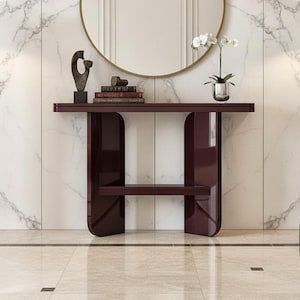 May include: A modern entryway scene featuring a burgundy console table with a lower shelf and rounded legs. A round gold-framed mirror hangs above the table, reflecting the room. A sculpture, books, and a potted orchid decorate the table.