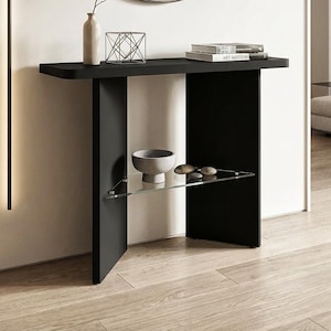 May include: A modern black console table with a minimalist design. It features a rectangular top, a lower glass shelf, and a geometric metal sculpture. A round mirror hangs above the table, reflecting the room's decor. A vase and books are also on the table.