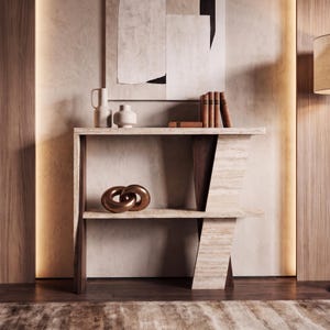 Op de afbeelding: Een console-tafel met twee niveaus, gemaakt van beige travertijnsteen en donker hout. Het bovenblad bevat twee keramische vazen en vier boeken. De onderste plank is voorzien van een metalen, bronskleurige sculptuur van in elkaar grijpende ringen.