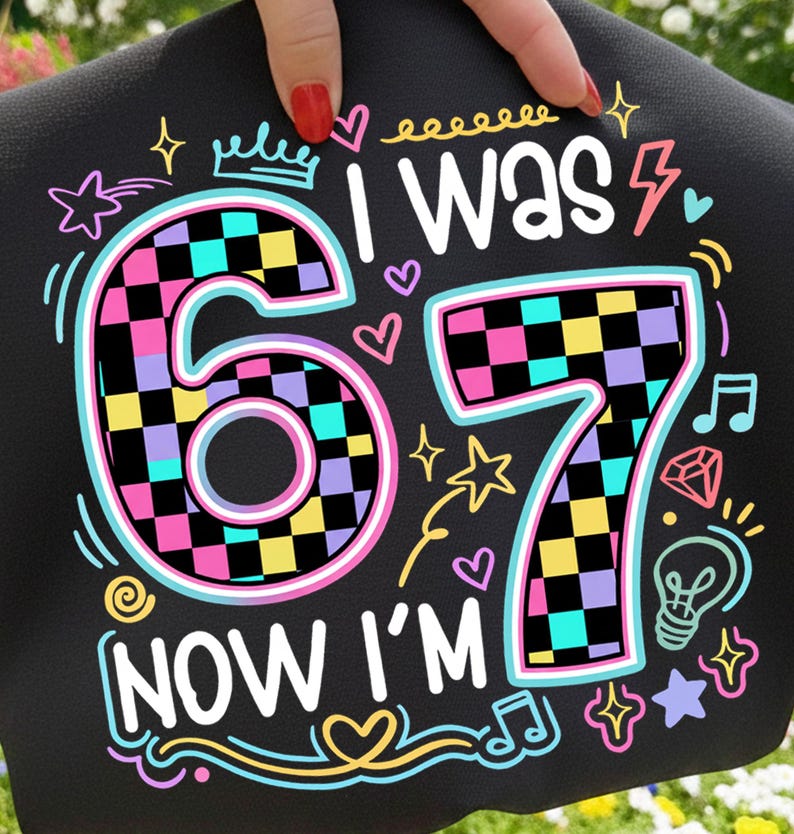 May include: Black fabric with the text "I WAS 67 NOW I'M" in white, with colorful accents. The numbers 6 and 7 are checkered with pink, yellow, black, and teal squares. The design includes stars, hearts, and musical notes.