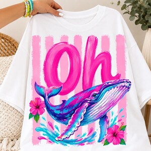 Pu&ograve; includere: Una t-shirt bianca con una stampa grafica di una balena blu e viola che nuota sopra spruzzi d'acqua. Il design include la parola Oh in grandi lettere corsive rosa su strisce verticali rosa, accentuato da due fiori di ibisco rosa brillante.