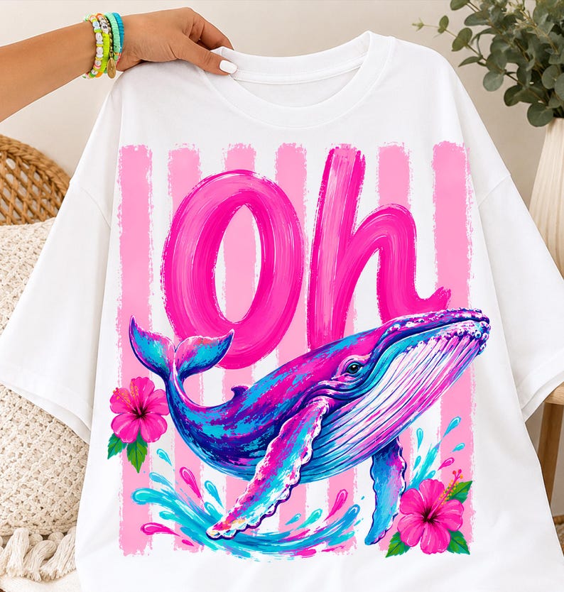 Pu&ograve; includere: Una t-shirt bianca con una stampa grafica di una balena blu e viola che nuota sopra spruzzi d'acqua. Il design include la parola Oh in grandi lettere corsive rosa su strisce verticali rosa, accentuato da due fiori di ibisco rosa brillante.